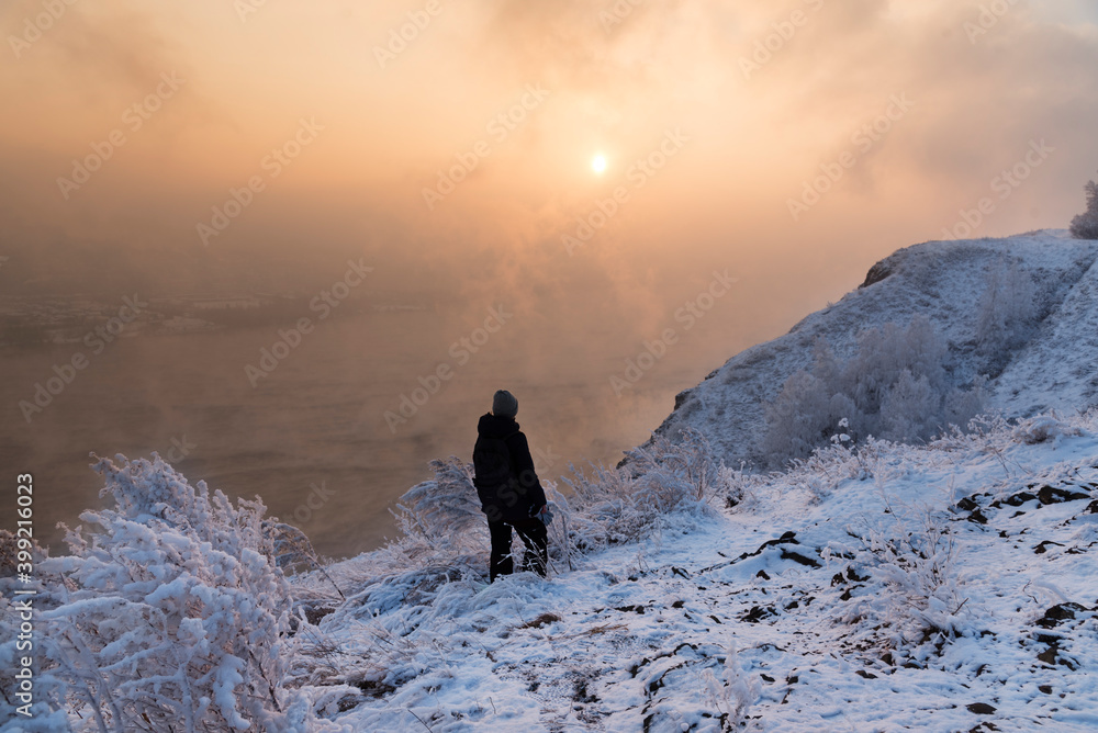 A girl admires the river sunset in winter, standing on a rock. Beautiful, wintry, landscape, river without ice, sunset