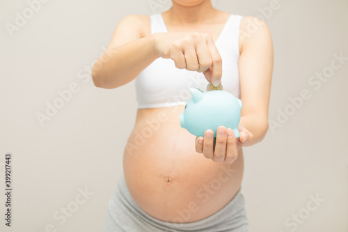Money saving, donation, economizing, pregnancy, reservation, finance, investment and life insurance concept - young beautiful pregnant woman dropping golden coin into piggy bank glass for saving mone