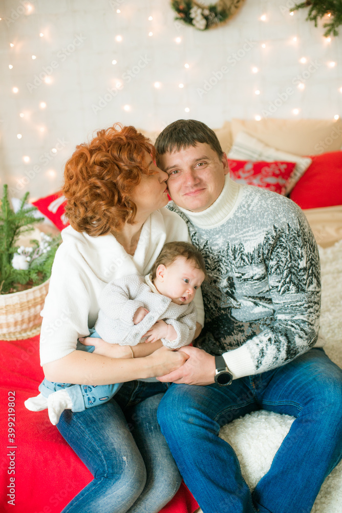 A young family in warm sweaters with a baby daughter in a Christmas bedroom interior decorated with red blankets, pillows, garlands and green pine needles. Family and Children. Christmas mood
