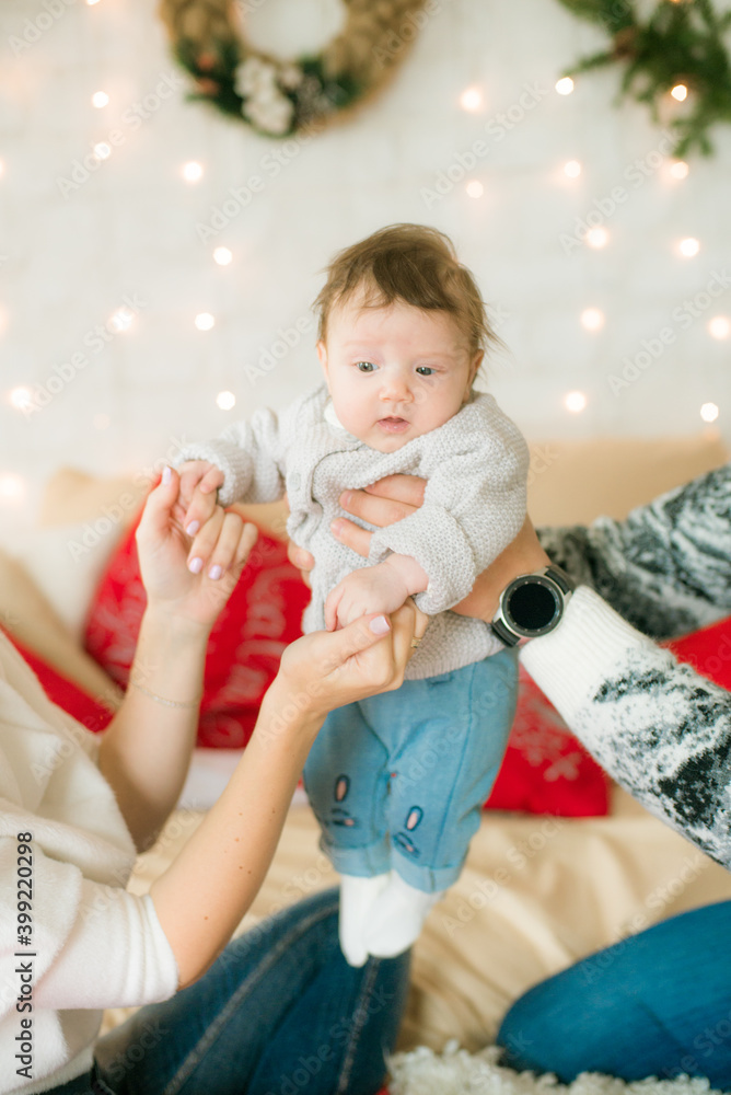 Fototapeta premium A young family in warm sweaters with a baby daughter in a Christmas bedroom interior decorated with red blankets, pillows, garlands and green pine needles. Family and Children. Christmas mood 