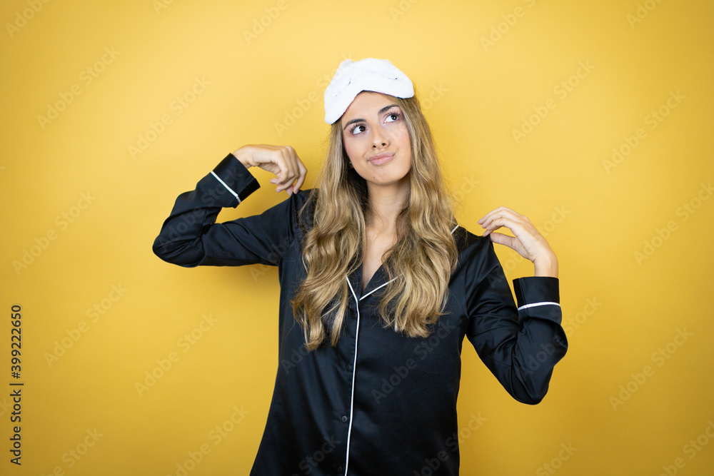 Young pretty woman wearing sleep mask and pajamas over isolated yellow background holding her t-shirt with a successful expression and cockiness Young pretty woman wearing sleep mask and pajamas over isolated yellow background holding her t-shirt with a successful expression and cockiness