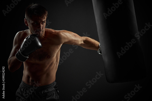 muscular boxer in black boxing gloves punching in boxing bag on dark background