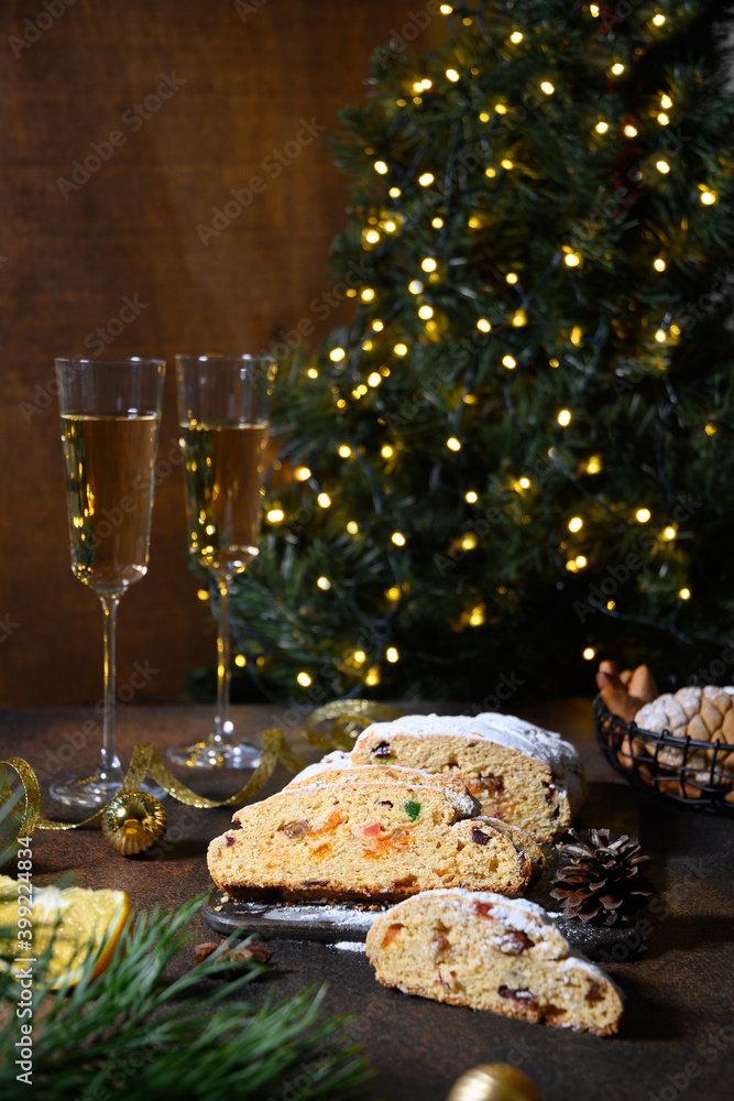 Homemade festive stollen and two glasses of sparkling wine on dark table in front of the Christmas tree. Close up. Holiday Xmas food.