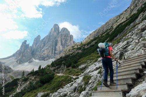 Woman with big backpack and sticks, walking up a wooden stairs in high Italian Dolomites. There are many sharp peaks in front of her. She is going up. There are a few trees around. Sunny day. Outdoor