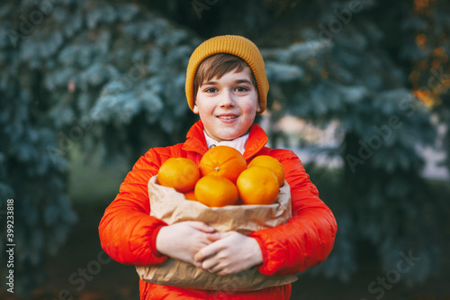 A boy in a bright orange jacket and a yellow hat holds a large package with oranges in his hands against the background of a Christmas tree with a surprised face. Preparing for Christmas, New Year