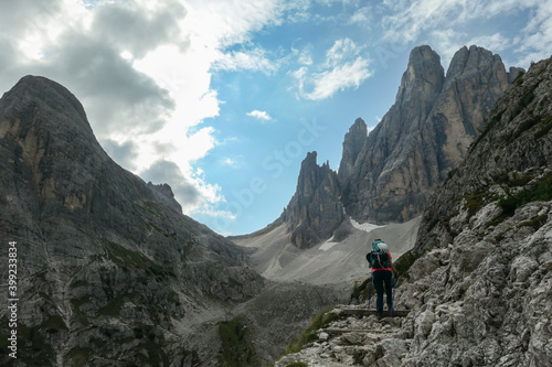 A woman with big backpack and sticks, hiking in high Italian Dolomites. There are many sharp peaks in front of her. She is going up. Lots of lose stones and landslides. Sunny day. Outdoor exercising © Chris
