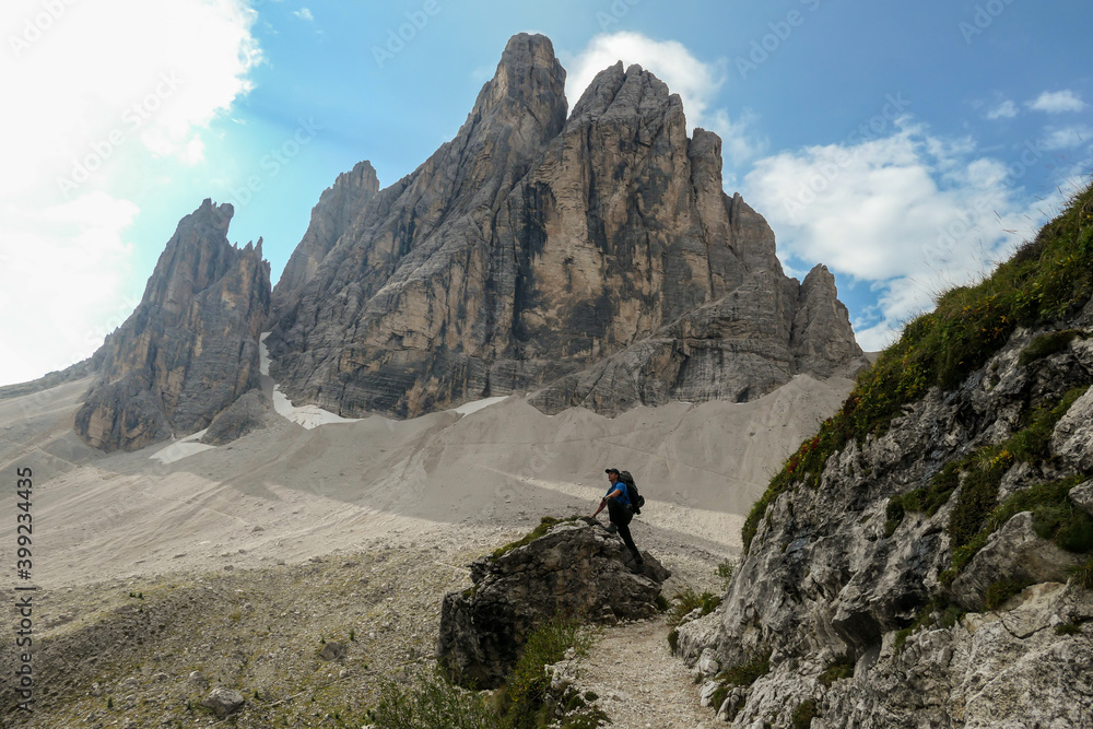 Man with big backpack and sticks, hiking in high Italian Dolomites. There are many sharp peaks in front of him. He is standing on a big boulder. Lots of lose stones and landslides. Sunny day. Outdoor