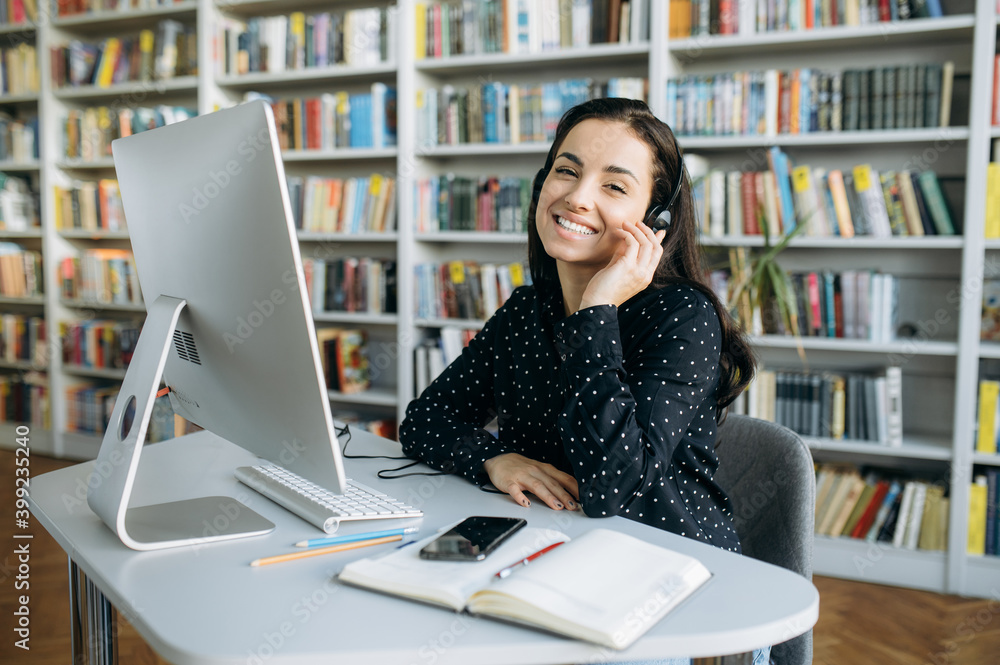 Remote work. Happy young pretty woman, call center worker, teacher or manager, wearing a headset, looks at the camera with smile while sitting at her workplace Remote work. Happy young pretty woman, call center worker, teacher or manager, wearing a headset, looks at the camera with smile while sitting at her workplace