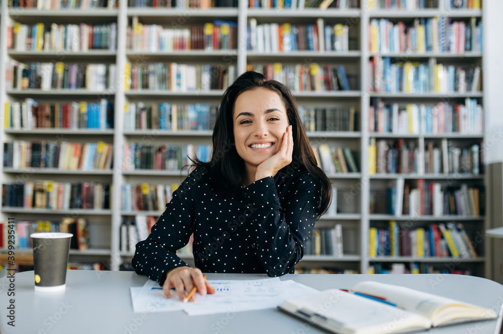 Portrait of an attractive young woman, student, teacher or business woman, looking with a smile directly at the camera sitting on her workplace indoors Portrait of an attractive young woman, student, teacher or business woman, looking with a smile directly at the camera sitting on her workplace indoors
