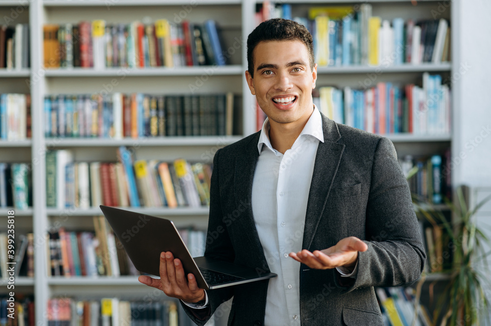 Portrait of friendly attractive hispanic man looking at camera with a pleasant smile and holding laptop in hand, indoors Portrait of friendly attractive hispanic man looking at camera with a pleasant smile and holding laptop in hand, indoors