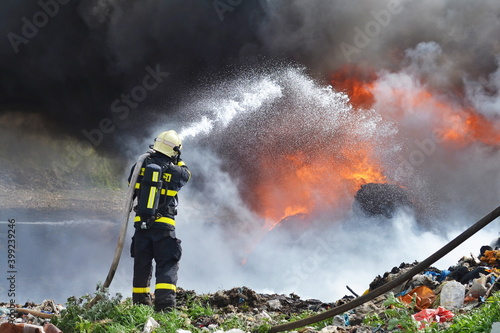 Photo A fireman extinguishes huge landfill fire with flames and black smoke in the bac
