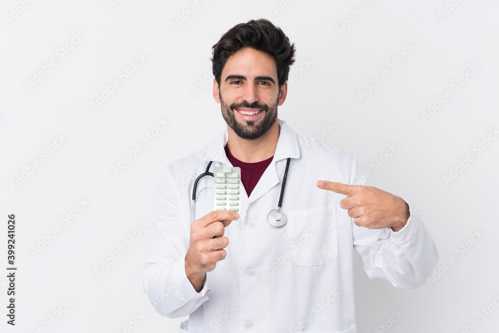 Young handsome man with beard over isolated white background wearing a doctor gown and holding pills
