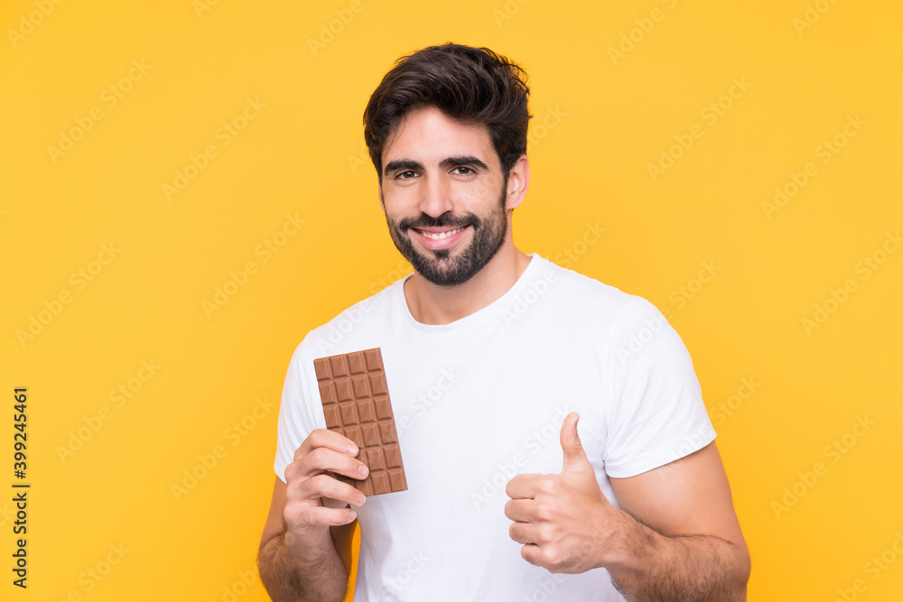 Young handsome man with beard over isolated yellow background taking a chocolate tablet and with thumb up Young handsome man with beard over isolated yellow background taking a chocolate tablet and with thumb up