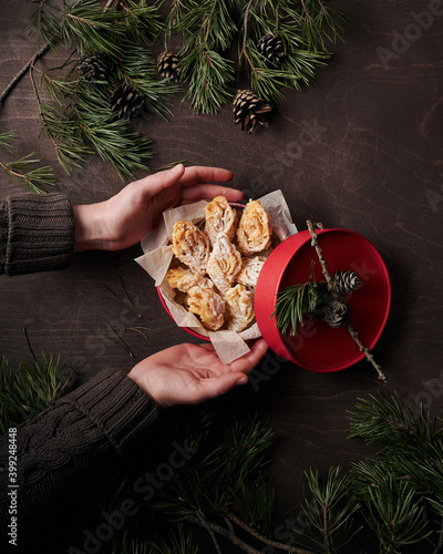 Opening a gift. Round box with pastry among pine branches and cones on dark brown wooden table, young woman’s hands holding it from the left, have just opened the box photo