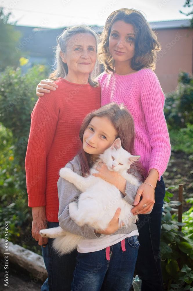 Senior mother with gray hair with her adult daughter and little granddaughter with white cat are looking at the camera in the garden and hugging each other during sunny day outdoors Senior mother with gray hair with her adult daughter and little granddaughter with white cat are looking at the camera in the garden and hugging each other during sunny day outdoors