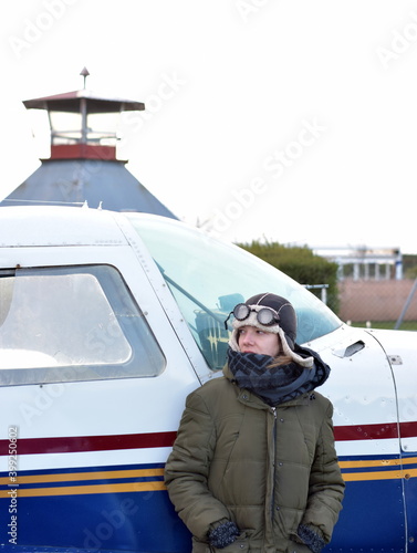 Woman with cap and aviator glasses with green coat and blue scarf leaning on the plane, while looking at the tail of the plane. In the background the control tower