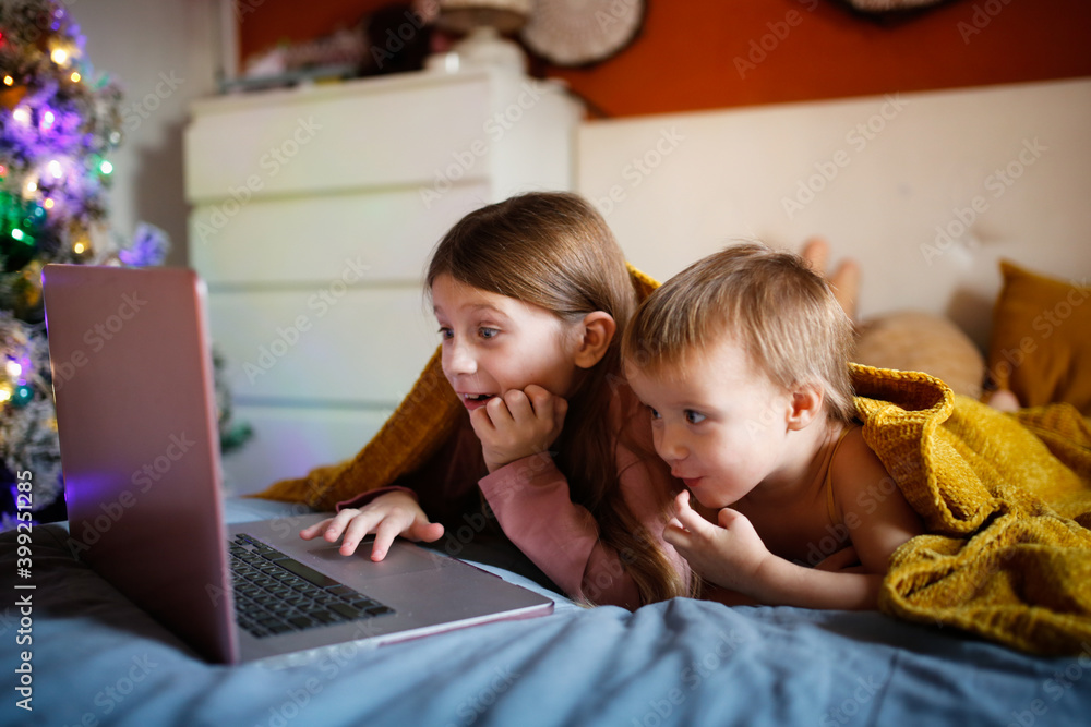 Girls children in bedroom are surprised in delight looking at laptop screen, concept of Christmas sales and interesting cartoons. Children sisters on bed with blanket lying looking at laptop Girls children in bedroom are surprised in delight looking at laptop screen, concept of Christmas sales and interesting cartoons. Children sisters on bed with blanket lying looking at laptop