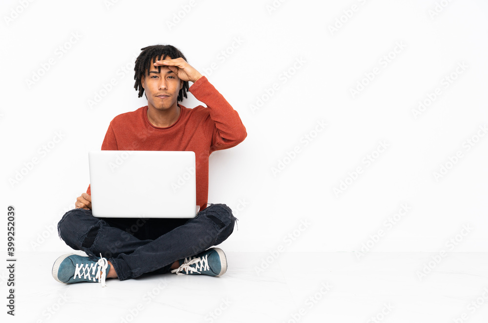 Young african american man sitting on the floor and working with his laptop looking far away with hand to look something Young african american man sitting on the floor and working with his laptop looking far away with hand to look something