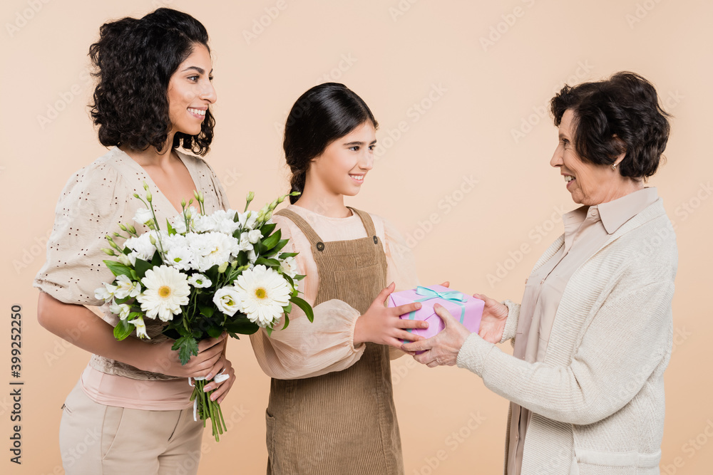 Smiling hispanic girl giving present to grandmother near mother with flowers isolated on beige, three generations of women Smiling hispanic girl giving present to grandmother near mother with flowers isolated on beige, three generations of women