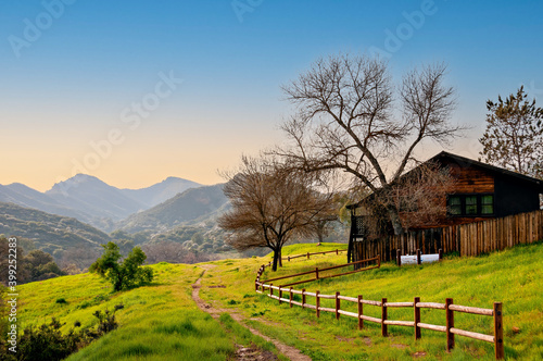 Serene southern California rural landscape with oak trees, fenced wooden hut on green meadow and mountains background. photo
