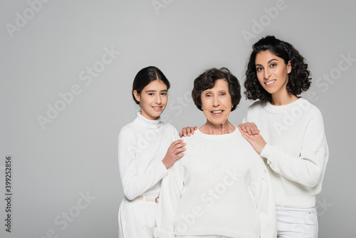 Hispanic woman and daughter embracing smiling grandmother isolated on grey, three generations of women