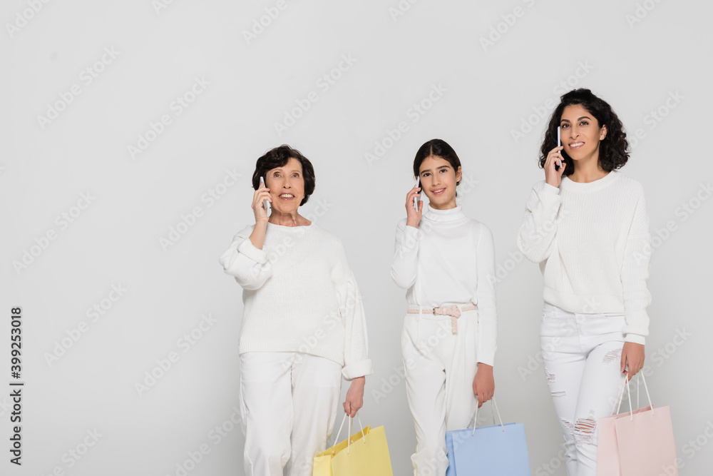 Cheerful hispanic women and kid talking on smartphones and holding shopping bags isolated on grey, three generations of women Cheerful hispanic women and kid talking on smartphones and holding shopping bags isolated on grey, three generations of women