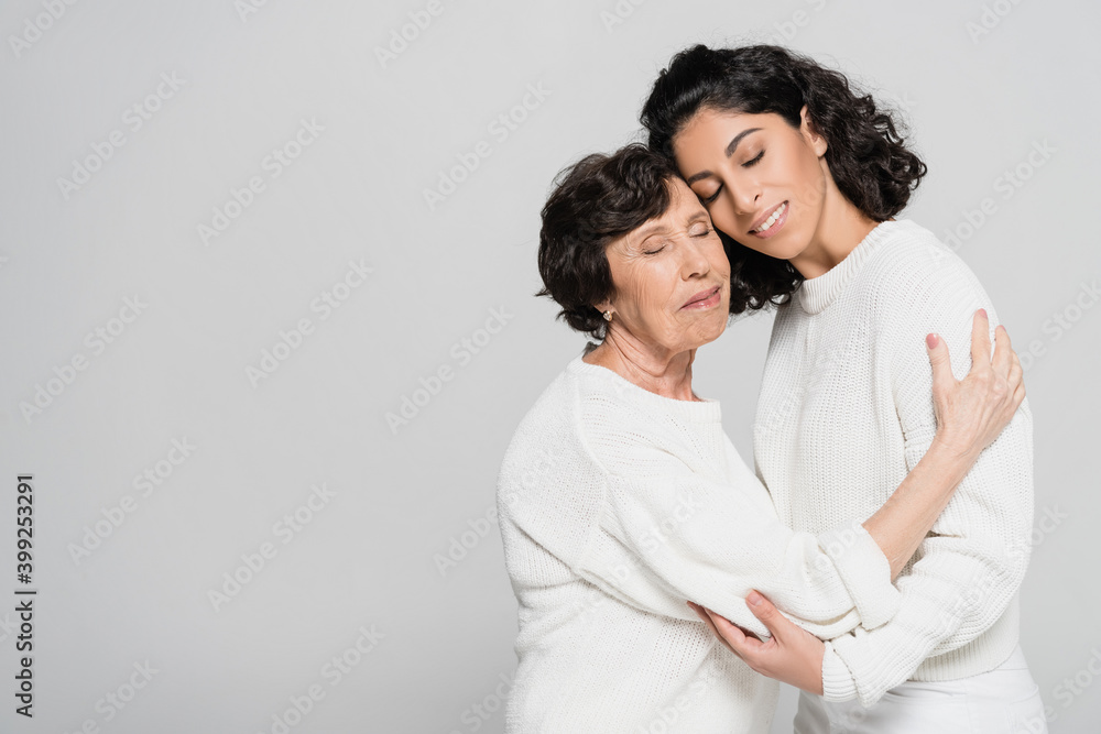 Senior woman embracing hispanic daughter with closed eyes isolated on grey, two generations of women Senior woman embracing hispanic daughter with closed eyes isolated on grey, two generations of women
