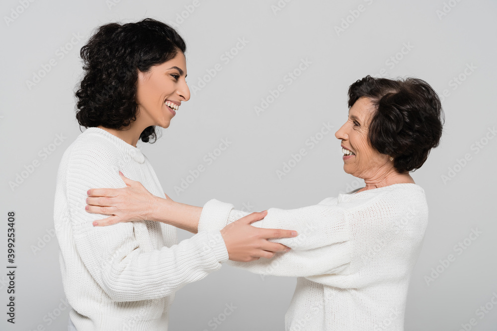 Side view of cheerful hispanic woman hugging elderly mother isolated on grey, two generations of women Side view of cheerful hispanic woman hugging elderly mother isolated on grey, two generations of women