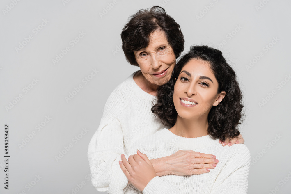 Hispanic woman smiling while touching hand of senior mother isolated on grey, two generations of women Hispanic woman smiling while touching hand of senior mother isolated on grey, two generations of women