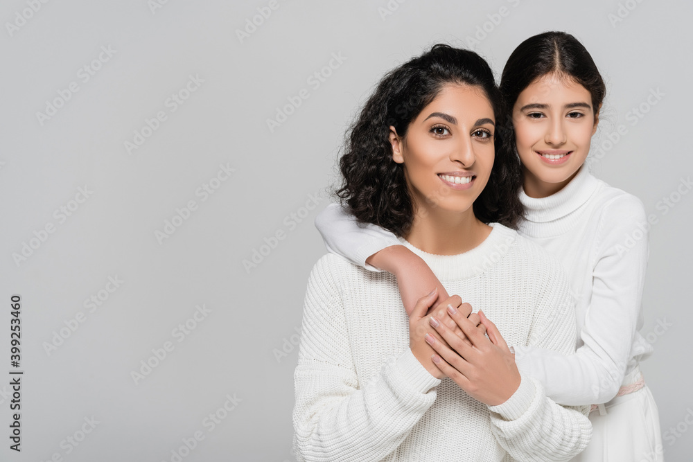 Cheerful hispanic child embracing mother and looking at camera isolated on grey, two generations of women Cheerful hispanic child embracing mother and looking at camera isolated on grey, two generations of women