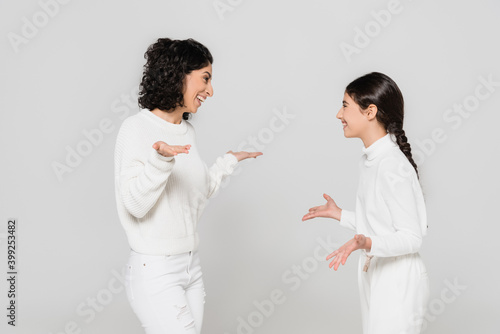 Smiling hispanic woman pointing with hands near cheerful daughter isolated on grey, two generations of women