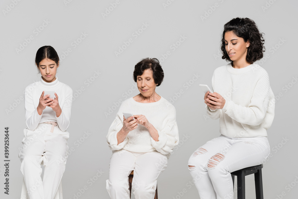 Hispanic women and kid using smartphones on chairs isolated on grey, three generations of women Hispanic women and kid using smartphones on chairs isolated on grey, three generations of women