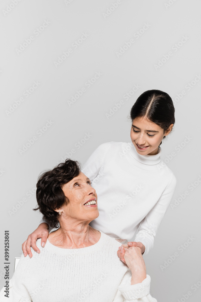 Smiling hispanic girl holding hand and smiling at grandmother isolated on grey, two generations of women Smiling hispanic girl holding hand and smiling at grandmother isolated on grey, two generations of women