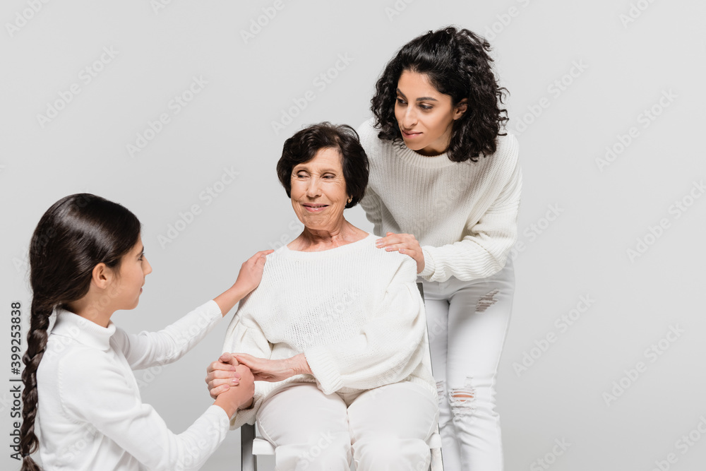 Hispanic woman and child holding hand of granny on chair isolated on grey, three generations of women Hispanic woman and child holding hand of granny on chair isolated on grey, three generations of women