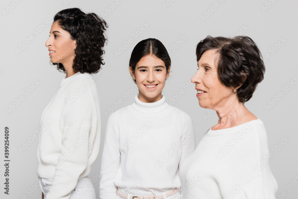 Smiling hispanic girl looking at camera near mother and granny isolated on grey, three generations of women Smiling hispanic girl looking at camera near mother and granny isolated on grey, three generations of women