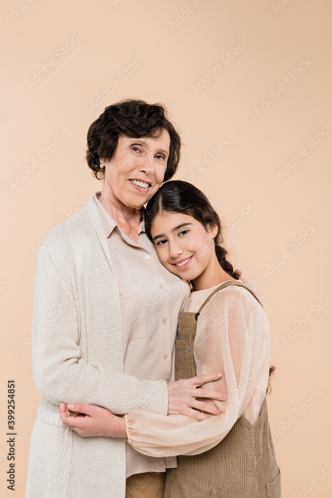 Hispanic woman smiling at camera while embracing granddaughter isolated on beige, two generations of women Hispanic woman smiling at camera while embracing granddaughter isolated on beige, two generations of women