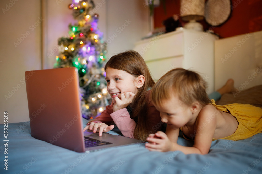 Girls children in bedroom are looking at screen near Christmas tree in real interior, concept of Christmas sales and interesting cartoons. siblings on the bed with blanket lying looking laptop Girls children in bedroom are looking at screen near Christmas tree in real interior, concept of Christmas sales and interesting cartoons. siblings on the bed with blanket lying looking laptop