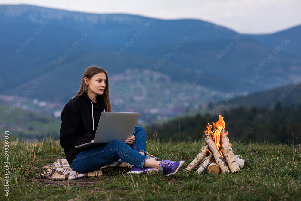 young female freelancer working on laptop in the mountains in the evening. Tourist girl sitting near campfire and having fun. Copy space.
