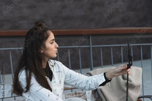 Young hipster brunette taking a selfie or video with front camera on mobile phone, student with a funny video call and laughing in the park during a break in college, wifi in public places
