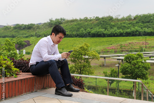 Asian college male student sit on red bricks while using mobile phone in beautiful university campus with grass, tree and red brick walls.