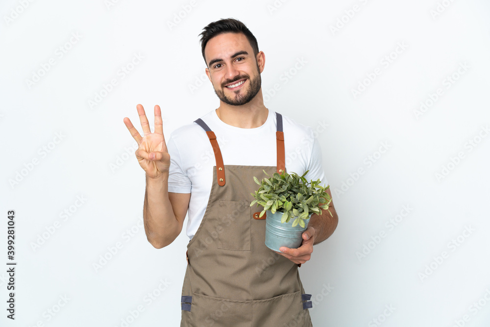 Gardener man holding a plant isolated on white background happy and counting three with fingers Gardener man holding a plant isolated on white background happy and counting three with fingers
