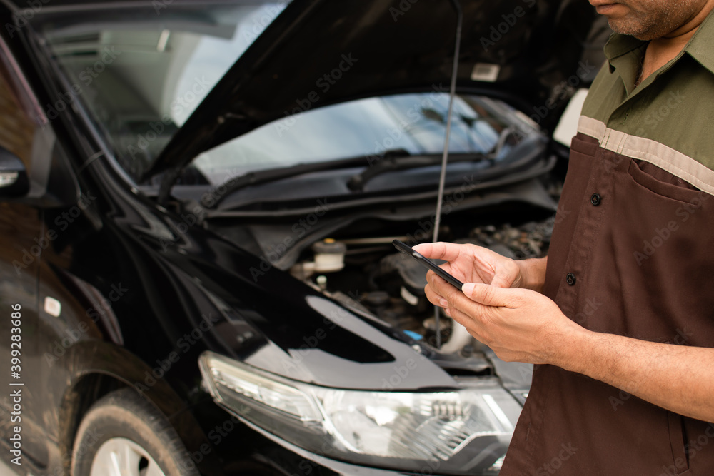 Close-up, A male technician stands holding a smartphone to check the operation of the black car's engine in the garage. The engine of a car does not start. A man using a smartphone. Blurred background Close-up, A male technician stands holding a smartphone to check the operation of the black car's engine in the garage. The engine of a car does not start. A man using a smartphone. Blurred background