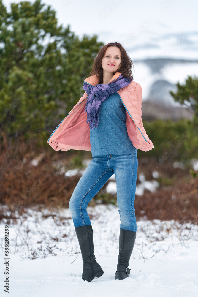 Young woman stands in winter forest near evergreen coniferous tree, looking at camera. Cute brunette woman with long hair, dressed in beige jacket with scarf, blue jeans, high-necked demi-season boots Young woman stands in winter forest near evergreen coniferous tree, looking at camera. Cute brunette woman with long hair, dressed in beige jacket with scarf, blue jeans, high-necked demi-season boots