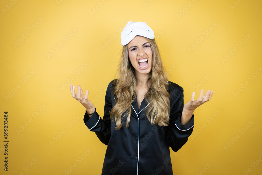 Young pretty woman wearing sleep mask and pajamas over isolated yellow background crazy and mad shouting and yelling with aggressive expression and arms raised. Frustration concept. Young pretty woman wearing sleep mask and pajamas over isolated yellow background crazy and mad shouting and yelling with aggressive expression and arms raised. Frustration concept.