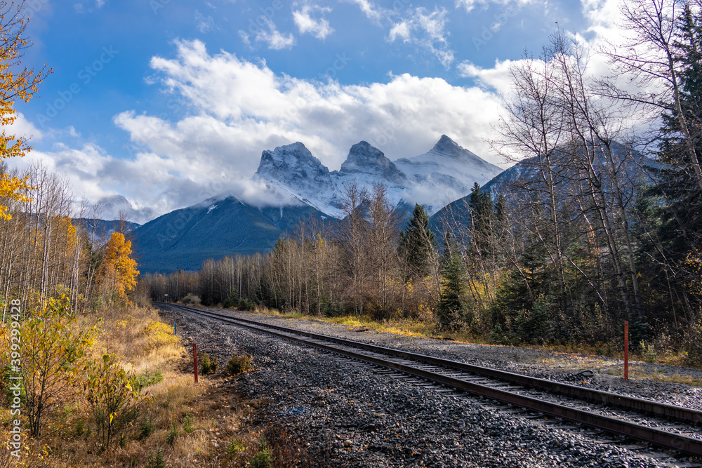 Railway scenery in autumn season, blue sky with white clouds and snow capped The Three Sisters trio of peaks in the background. Canmore, Alberta, Canada. Railway scenery in autumn season, blue sky with white clouds and snow capped The Three Sisters trio of peaks in the background. Canmore, Alberta, Canada.