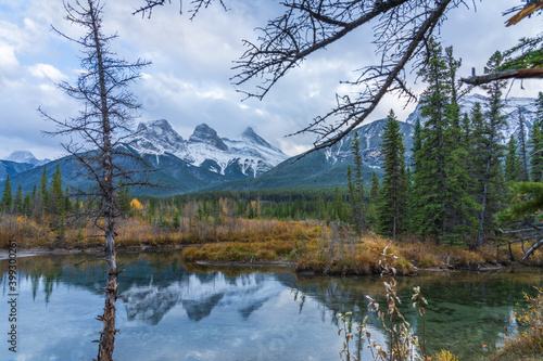 Fototapeta Naklejka Na Ścianę i Meble -  Snow capped The Three Sisters trio peaks mountain with blue sky and white clouds reflect on water surface in autumn. Beautiful natural scenery landscape at Canmore, Canadian Rockies, Alberta, Canada.