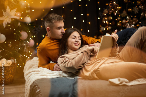 loving couple is celebrating Christmas at home. couple in love is hugging and reading book near Christmas tree