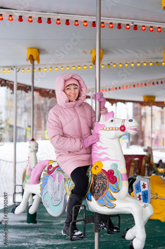 A young woman in a pink down jacket rides in winter in a city park on a children's carousel