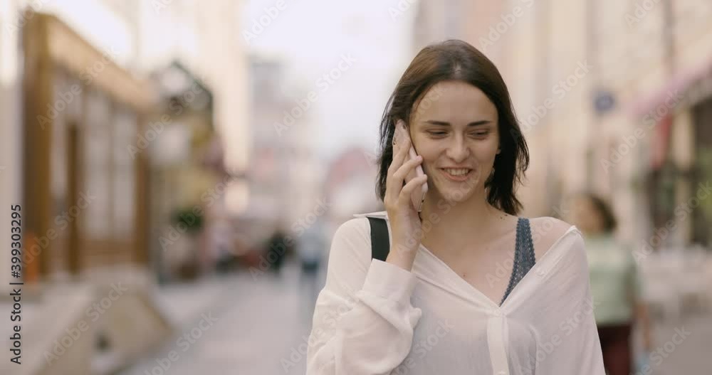 Young woman walking in the street, talking nicely on the phone and smiles enjoys a conversation with her beloved or friend Young woman walking in the street, talking nicely on the phone and smiles enjoys a conversation with her beloved or friend