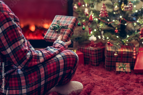 Woman in pajamas with christmas gift box in hand sitting on fluffy plaid near fireplace and christmas tree.